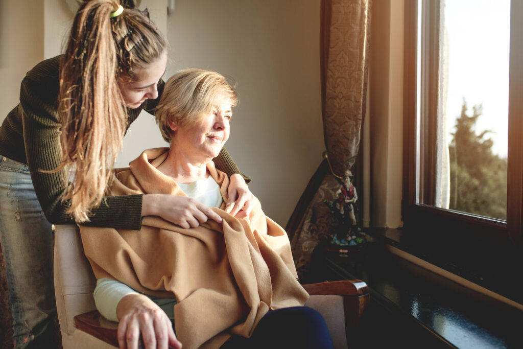 Younger woman wrapping shawl around senior woman sitting in chair looking out window